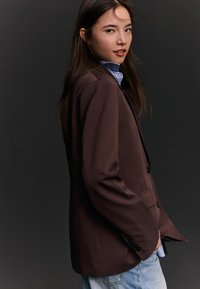 Mlada ženska z dolgimi temnimi lasmi, oblečena v rjavo srajco in svetlo modro srajco, ki pozira iz strani na temno sivi ozadju.