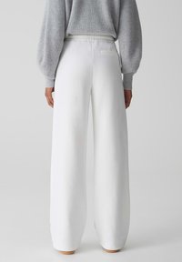 Weiße, weit geschnittene Sweatpants mit einer weichen Textur, ausgestattet mit einem hohen Bund und einer Gesäßtasche, kombiniert mit einem hellgrauen Strickpullover.