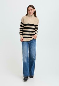 Beige und schwarz gestreifter Strickpullover mit hohem Kragen und Reißverschluss, kombiniert mit weit geschnittenen blauen Jeans und schwarzen Schuhen, auf einem hellen Hintergrund.