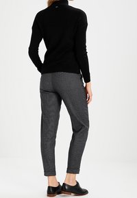 Schwarzer Rollkragenpullover kombiniert mit grauen strukturierten Hosen, die umgeschlagene Säume und schwarze Schnürschuhe aufweisen. Einfarbiger Hintergrund.