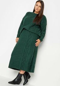 Conjunto de suéter verde de punto y falda a juego, que presenta un patrón de trenzado texturizado. Llevado con botas negras hasta el tobillo.