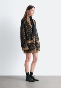 Übergroßer Cardigan mit Leopardenmuster in Braun und Schwarz, aus einem flauschigen Material, mit zwei Fronttaschen und Knopfverschluss. Schwarzes Hemd und Schuhe.