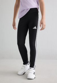 Kind trägt schwarze Adidas Leggings mit weißen Streifen und weiße Adidas Sneaker, steht vor einem schlichten Hintergrund.