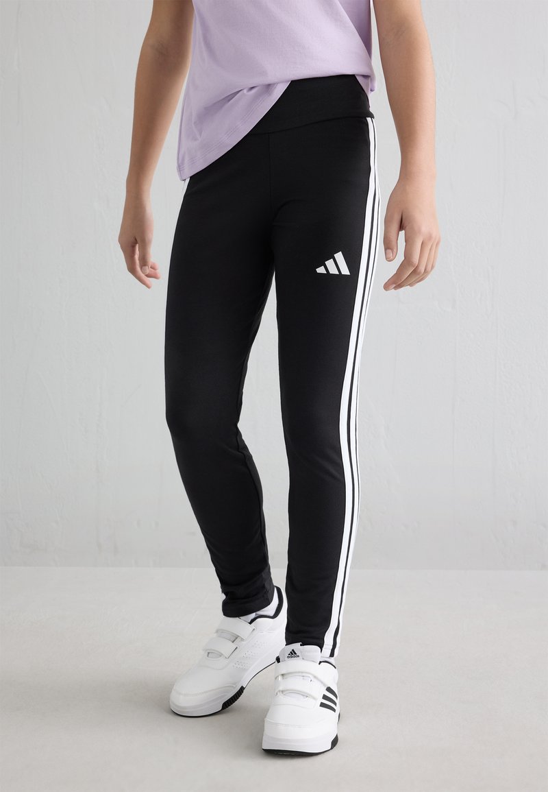 Kind trägt schwarze Adidas Leggings mit weißen Streifen und weiße Adidas Sneaker, steht vor einem schlichten Hintergrund.