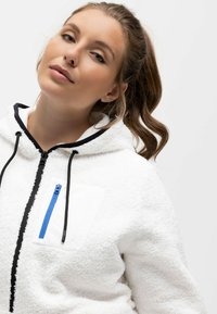 Sudadera de felpa blanca con un acabado texturizado, que presenta un bolsillo con cremallera azul, cordones negros y un ribete negro en contraste alrededor de la capucha.