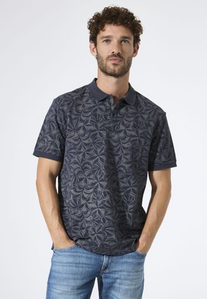 Man met krullend haar en baard, draagt een donkerblauw poloshirt met bloemmotief en lichtblauwe spijkerbroek, handen in zijn zakken, tegen een witte achtergrond.