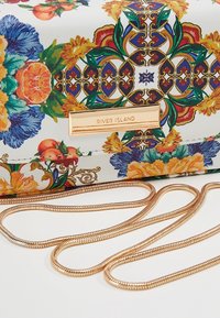 Květinová kabelka s bílým základem, zlatým řetízkovým popruhem a zlatou logovou destičkou, se zářivými modrými, oranžovými a zelenými detaily.