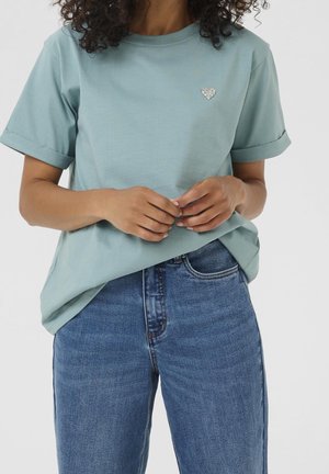 Kobieta w jasnoniebieskim T-shircie z podwiniętymi rękawami, z małą broszką w kształcie serca z kryształków i niebieskich dżinsach, ręce złożone na talii.