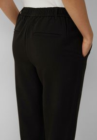 Pantalons noirs avec une taille élastique, texture lisse, deux poches arrière et une coupe ajustée, vus de derrière.