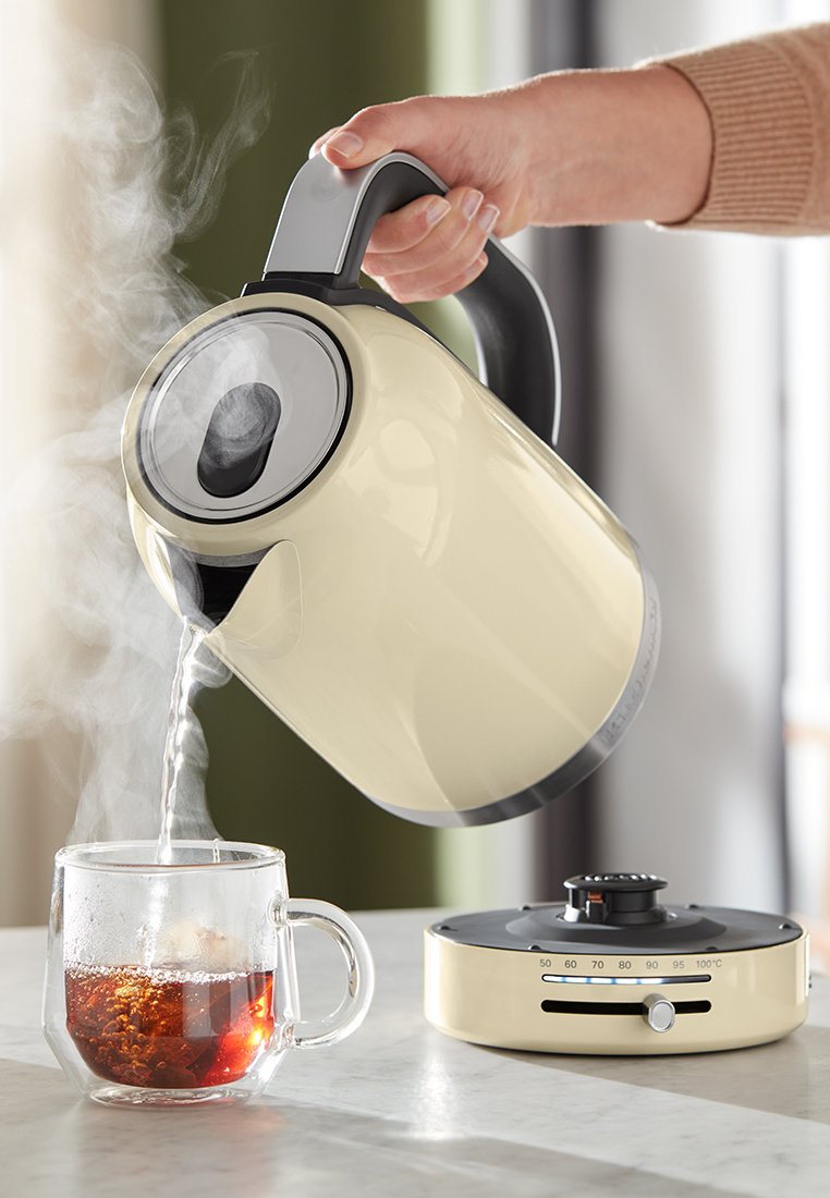 Bouilloire électrique crème et noire, avec un corps arrondi et un bec verseur. L'eau se verse dans une tasse en verre transparent, avec de la vapeur s'élevant visiblement au-dessus.