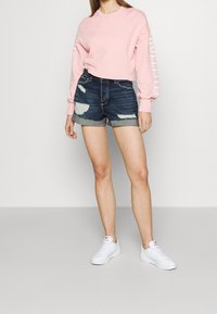 Lichtroze cropped sweatshirt met lange mouwen en witte tekst, gecombineerd met donkerblauwe distressed denim shorts en witte sneakers.