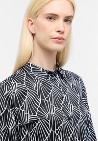 Chemise à motifs noir et blanc avec col, présentant des lignes géométriques sur un tissu lisse, fermeture à boutons et un design ajusté.