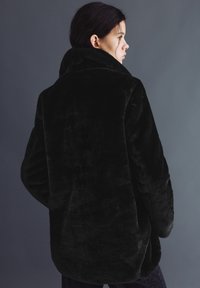 Tânără femeie văzută din profil purtând o haină neagră de blană artificială plușată, pe un fundal gri simplu.