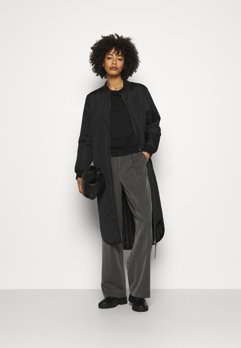 Manteau long noir matelassé avec col côtelé, associé à un haut noir et un pantalon gris à jambe large. Pour compléter le look, un petit sac noir.