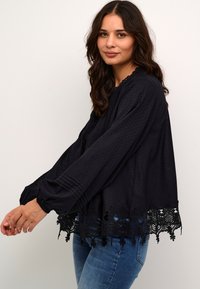 Melna blūze, kas izgatavota no teksturētas auduma ar punktiem, izšūta mežģīņu apakšmala un garām piedurknēm ar drapējuma detaļām. Kombinēta ar džinsiem.