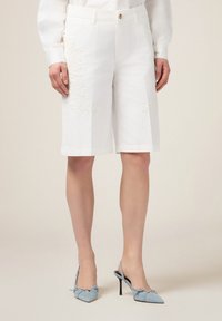Pantalones cortos blancos hasta la rodilla con detalle floral bordado en el lado izquierdo, con cierre de botón y bolsillos. Textura de tela suave.