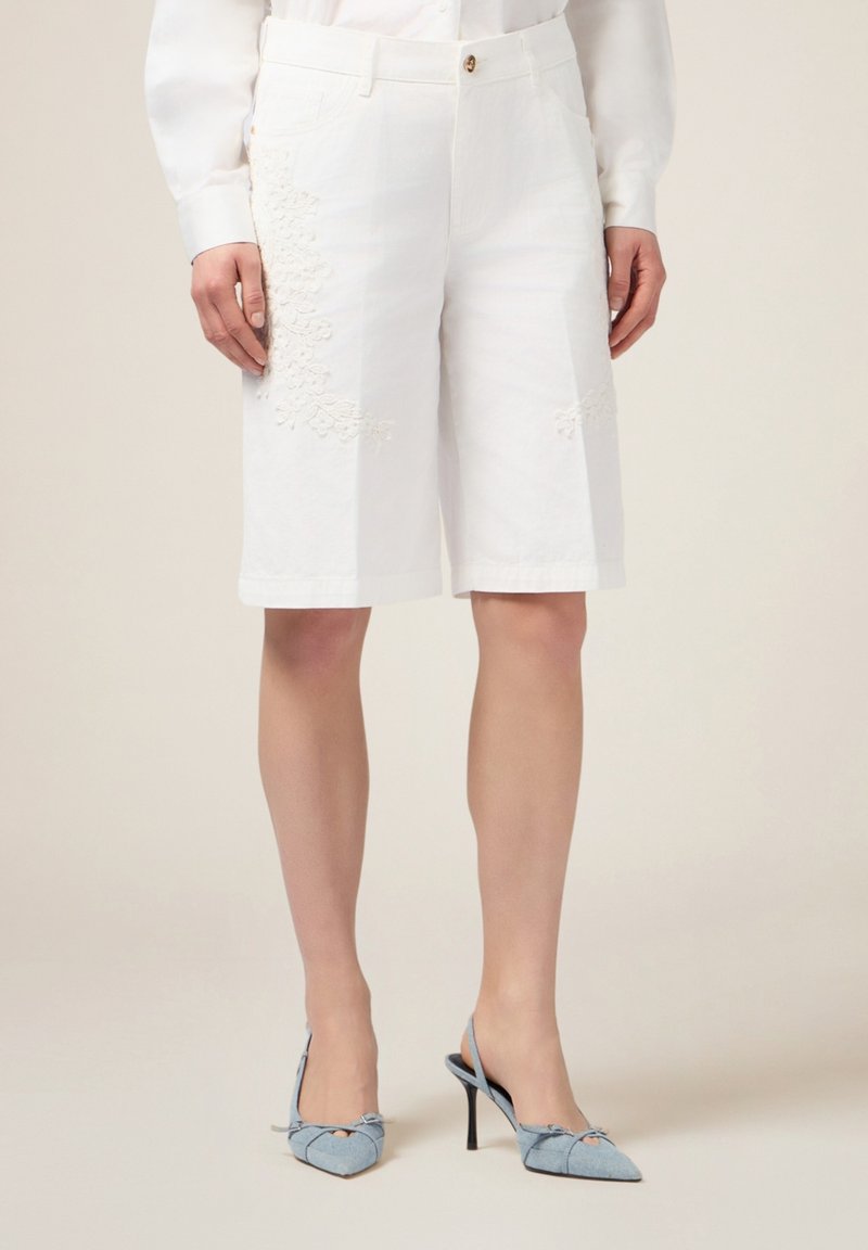 Pantalones cortos blancos hasta la rodilla con detalle floral bordado en el lado izquierdo, con cierre de botón y bolsillos. Textura de tela suave.