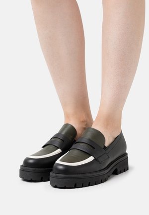 Chaussures en cuir noir et olive foncé avec une semelle épaisse, présentant une touche de blanc et deux sangles noires sur le dessus.