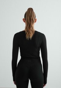Musta pitkähihainen paita, jossa on tiukka muotoilu, yhdistettynä korkeavyötäröisiin mustiin legginsseihin. Silkkinen pinta, vähäinen brändäys vyötärölinjassa.