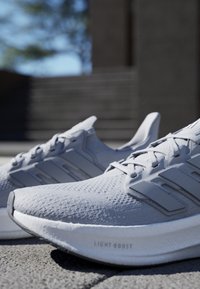 Svijetlosive atletske tenisice s teksturiranim pletenim gornjim dijelom, trostrukim oznakama brandiranja i debelim jastučićem označenim "LIGHT BOOST".