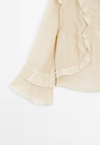 Blouse beige à manches longues avec poignets à volants et détail asymétrique à volants le long de l'avant et de la couture des manches.