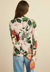 Long-sleeve floral shirt in light pink, featuring green leaves, red and white flowers. Textured fabric with a back seam detail. Paired with blue jeans.