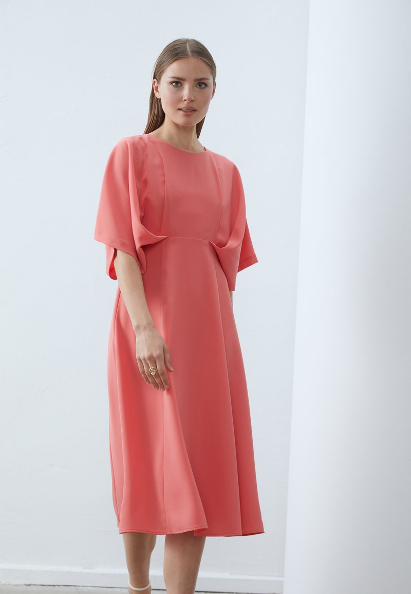 Robe midi corail avec des manches chauve-souris amples, en tissu lisse, avec un détail plissé à l'avant et une coupe décontractée, accompagnée d'une simple bague à la main.