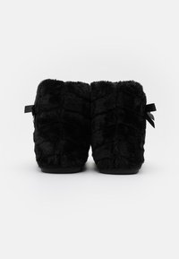 Schwarze, flauschige Hausschuhe mit strukturiertem Design und weicher Oberfläche. Mit einem Bandakzent an der Seite und flachen Sohlen.