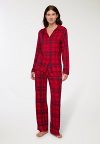 Completo pigiama a quadri rossi realizzato in tessuto morbido; presenta maniche lunghe, camicia con bottoni e pantaloni larghi abbinati con dettagli scuri.