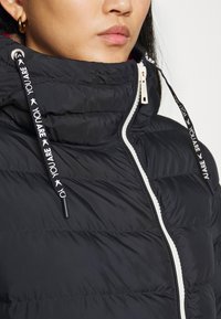 Czarna puchowa kurtka z wysokim kołnierzem, z białym zamkiem błyskawicznym i sznurkami do regulacji z tekstem "JESTEŚ". Teksturowany wzór pikowania na całej powierzchni.