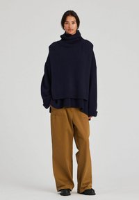 Kvinna som bär en oversize marinblå polotröja, lösa bruna byxor och svarta skor, stående mot en enkel ljus bakgrund.