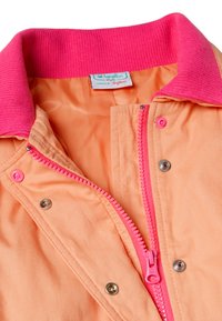 Jasnopomarańczowa kurtka z jaskraworóżowym prążkowanym kołnierzem, różowym zamkiem błyskawicznym i zatrzaskami, z widoczną wewnętrzną metką „Benetton”.