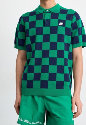 Mężczyzna ubrany w zielono-granatową, kraciastą koszulkę polo Nike oraz zielone szorty Nike Athletics na jednolitym tle.