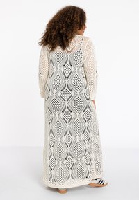Langes, cremefarbenes Häkelkleid mit einem schwarzen Diamantmuster. Volle Ärmel, offener Rücken und seitliche Schlitze für zusätzlichen Bewegungsspielraum.