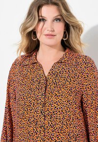 Camisa de botones de tela naranja con un patrón de print de leopardo que presenta acentos en negro y rosa, cuello clásico y placket de botones visible.