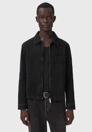 Jeune homme portant une veste noire zippée sur une chemise noire, jean foncé avec une ceinture noire, debout sur un fond clair uni.