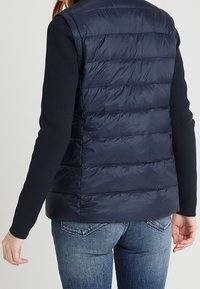 Tmavě modrá prošívaná vesta bez rukávů s měkkou texturou. Nošena přes top s dlouhým rukávem a v kombinaci s modrými džínami.