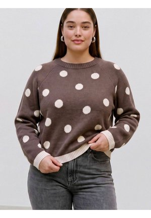 Femme aux longs cheveux bruns portant un pull marron à pois blancs et un jean gris, debout devant un fond clair uni.