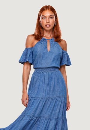 Donna con capelli rossi indossa un vestito blu scollato con scollo a goccia e gonna a balze su uno sfondo chiaro uniforme.
