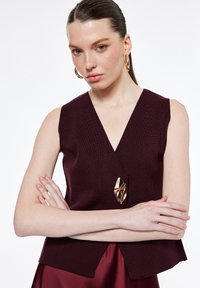 Vrouw met naar achteren gekamd haar, draagt gouden oorringen en een mouwloze donkerbordeauxrode top met een grote gouden gesp, armen gekruist.