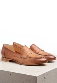 Mocasines de cuero marrón claro con punta redondeada, textura suave y detalles de costura mínimos. Tacón de madera bajo y diseño fácil de poner.