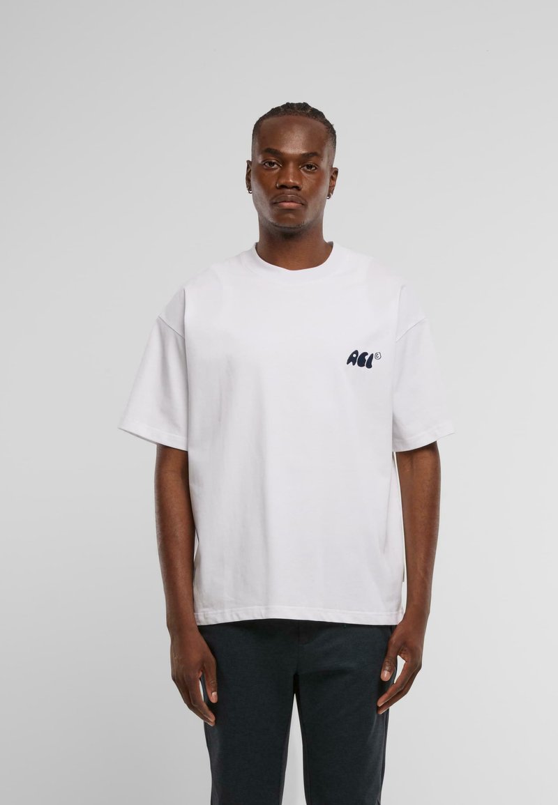 Biela bavlnená tričko s krátkymi rukávmi a okrúhlym výstrihom, s malým modrým potlačeným logom na ľavej hrudi. Model má na sebe tmavé nohavice.