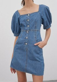 Denimmekko neliönmuotoisella kaula-aukolla, puffattuja lyhyitä hihoja ja etunappilistoilla. Mukana sivutaskut ja vyötäröltä muotoiltu malli. Vaalea sininen väri.
