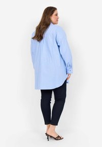 Světlomodrá oversized košile na knoflíky s dlouhými rukávy, zakulaceným lemem a skladem na zadní straně, spárovaná s tmavými skinny džíny a sandály na podpatku.