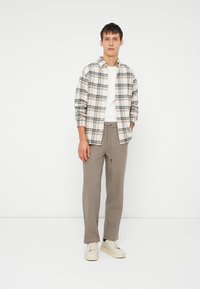 Jonge man die staat, gekleed in een beige geruite shirt over een witte t-shirt, grijze broek met trekkoord en witte sneakers tegen een witte achtergrond.