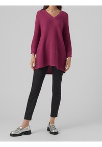 Femme portant un pull en maille ample magenta, un pantalon skinny noir et des mocassins argentés à semelles épaisses noires sur un fond gris uni.