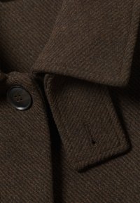 Brun ullblanding frakk detalj har en fishtail tekstur, knappelukking og klaffekrage. Stoffet har et mykt, varmt utseende.