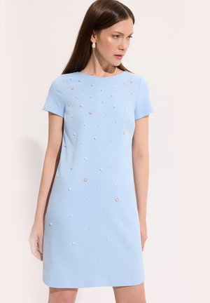 Kobieta ubrana w jasnoniebieską sukienkę z krótkim rękawem ozdobioną rozproszonymi małymi i średnimi perełkami oraz złotymi zdobieniami.