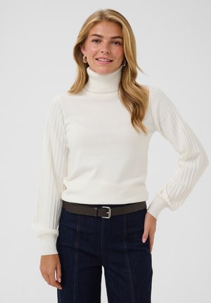 Femme aux cheveux blonds portant un pull à col roulé blanc avec des manches côtelées et un jean foncé avec une ceinture noire, souriante devant un fond uni.