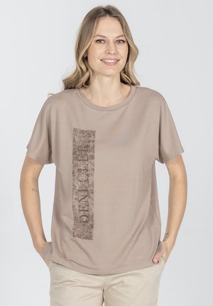 Kobieta o blond włosach w beżowej koszulce z pionowym kwiatowym wzorem i napisem "DEN.GE.R", stoi z rękami w kieszeniach jasnych spodni.